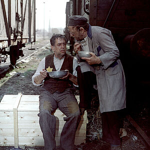 Ein Mann mit Judenstern sitzt zwischen zwei Zügen, isst und unterhält sich mit einem neben ihm stehenden Mann