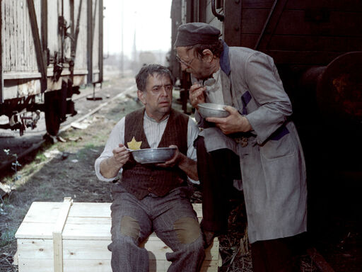 Ein Mann mit Judenstern sitzt zwischen zwei Zügen, isst und unterhält sich mit einem neben ihm stehenden Mann
