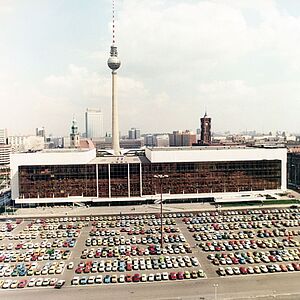 Das Bild zeigt den Palast der Republik. Der Parkplatz davor ist voll belegt. Im Hintergrund sind diverse Gebäude zu sehen, die ihn überragen. Allem vorran der Berliner Fernsehtrum und das Rote Rathaus.