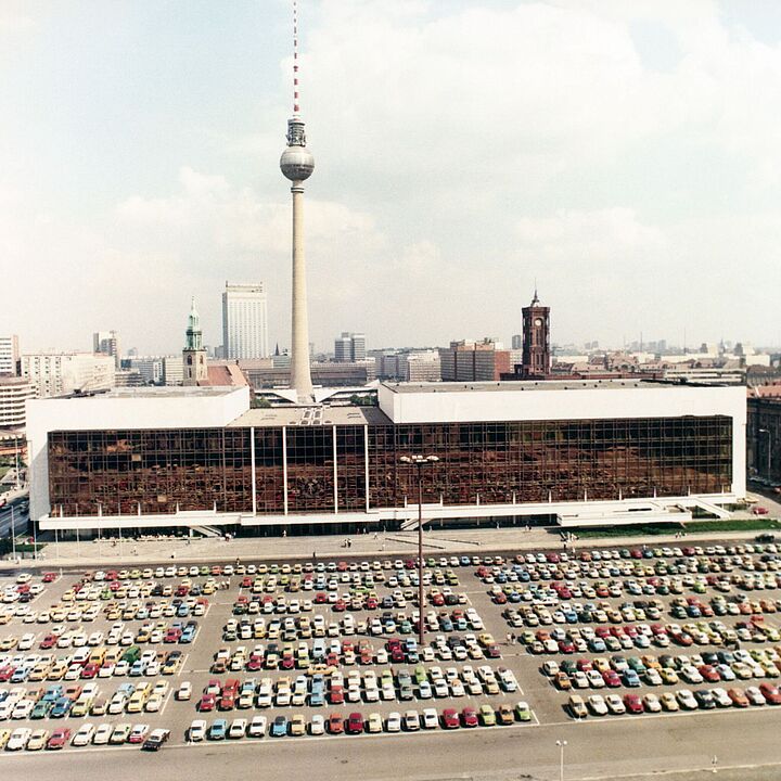 Das Bild zeigt den Palast der Republik. Der Parkplatz davor ist voll belegt. Im Hintergrund sind diverse Gebäude zu sehen, die ihn überragen. Allem vorran der Berliner Fernsehtrum und das Rote Rathaus.