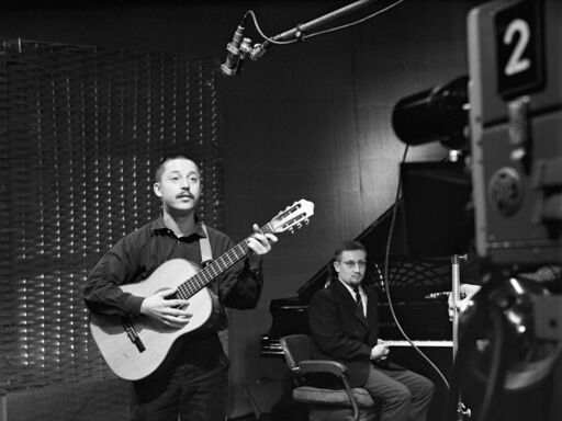Der junge Wolf Biermann steht mit Gitarre in einem Aufnahmestudio und singt, im Hintergrund sitzt ein Mann auf einem Klavierhocker