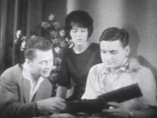 Mann zeigt stolz das Familienfotoalbum, in dem er bei der Verrichtung häuslicher Aufgaben zu sehen ist ... (Screenshot aus »Prisma« vom 11. Juli 1963) | Bildquelle: DRA Mann zeigt anderem Mann Fotoalbum, Frau sitzt ein bisschen im Hintergrund